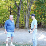Two men standing outdoors in a wooded area, engaged in conversation.