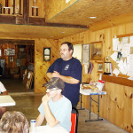 Two men in a wooden interior, one seated and one standing with arms crossed.