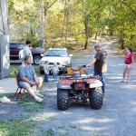 People relax and chat outdoors near an ATV and parked cars on a sunny day.
