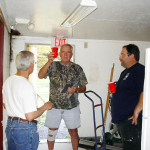Three men talking and holding tools in a room with cleaning supplies.