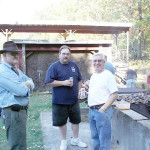 Three men standing outdoors near a barbecue grill, enjoying a casual gathering.