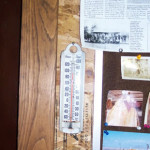 A vintage thermometer mounted on a wooden wall with photos and a newspaper clipping nearby.