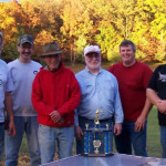 Five men posing outdoors with a trophy on a table in front of them.