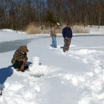 Three people ice fishing on a frozen lake surrounded by snow and trees.