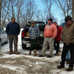 Four men standing outdoors in winter with a truck and leafless trees in the background.