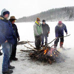 Five people warming by a campfire on a snowy landscape.