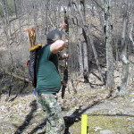 Person aiming a bow in a wooded area during daytime.