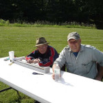 Two men sitting at a white outdoor table with drinks on a sunny day.