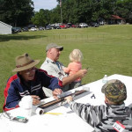 People sitting outside at a table with trees and cars in the background.
