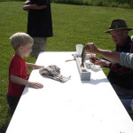 A young boy interacts with a man at an outdoor table on a sunny day.