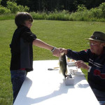 A young boy handing a fish to an older man outdoors at a table.