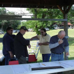 Four men standing under a canopy, engaging in conversation during an outdoor event.