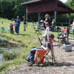 Children fishing by a small pond under a pavilion on a sunny day.