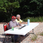 Two elderly men sitting at an outdoor table with papers and a drink.