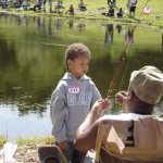 A young boy watches an older man fish by a pond in a park.