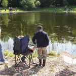 A child fishing by a calm lake with a folding chair nearby.