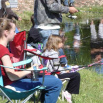 Two girls fishing by a pond with an adult nearby.