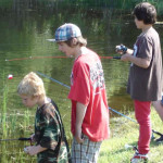 Three boys fishing by a calm lake on a sunny day.
