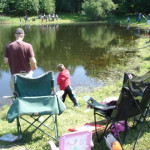 A family enjoying a sunny day by a pond with chairs and fishing gear.
