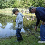 A child and an older man fishing by a lake on a sunny day.