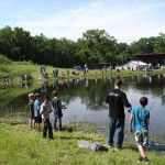 People fishing by a calm pond surrounded by trees and a small pavilion.