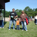 A family posing outdoors on a sunny day with others in the background.