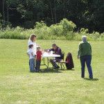 Children and adults gathered around a table outdoors in a grassy area.