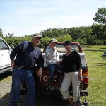 Three people posing cheerfully outdoors near a vehicle on a sunny day.