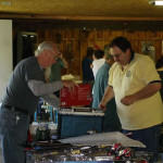 Two men at a table with tools and equipment, engaged in conversation.