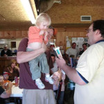 A man holds a toddler while another man offers a toy microphone indoors.