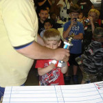 A child holding a microphone while standing near a whiteboard in a crowded room.