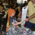 A child reads a paper while others watch around a recycling bin.