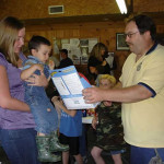 A man hands a certificate to a child held by a woman in a crowded indoor setting.