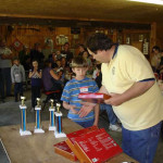 A man presents awards to a young boy in a crowded room.