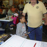 A young boy and an older man stand beside a large sheet with a grid at a community event.