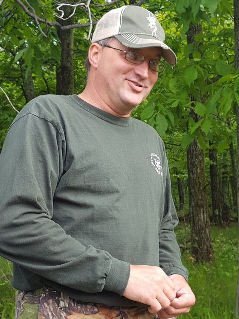 A man in a hat and glasses standing next to trees.